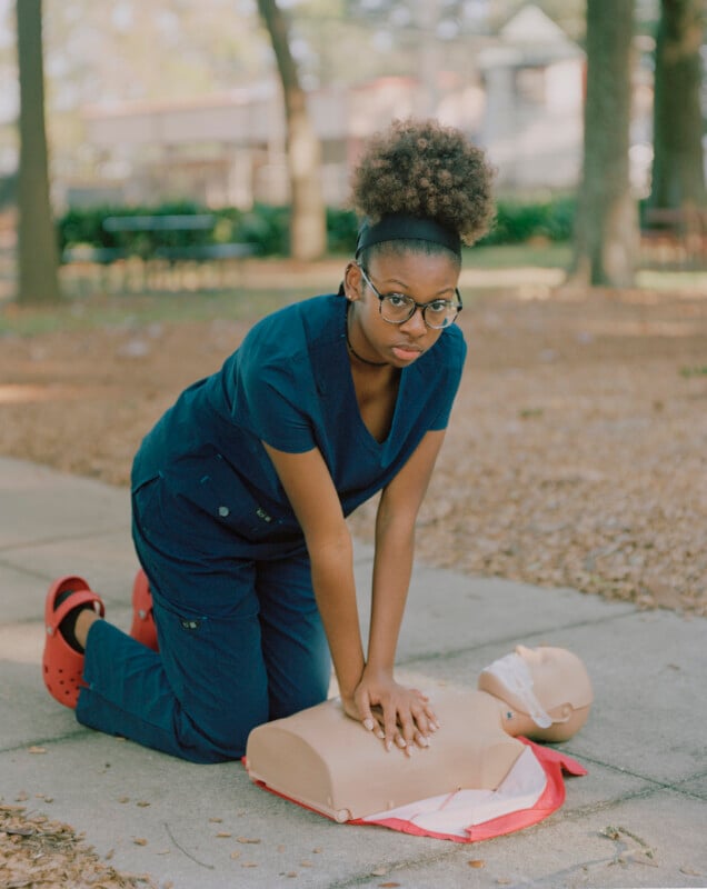 A young person in dark blue medical scrubs and red clogs kneels on a sidewalk, performing chest compressions on a CPR mannequin.