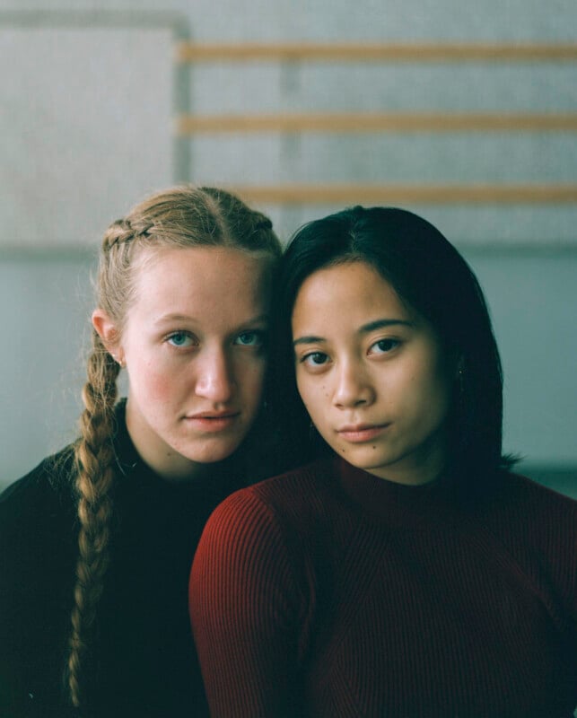 Two young women with serious expressions pose side-by-side, one with blonde braided hair in a black top and the other with dark hair in a red ribbed sweater.