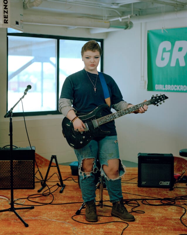 A person with short hair and dark makeup stands holding an electric guitar in a room with a microphone stand, amplifiers, and a green banner that reads "GR GIRLSROCKRO".