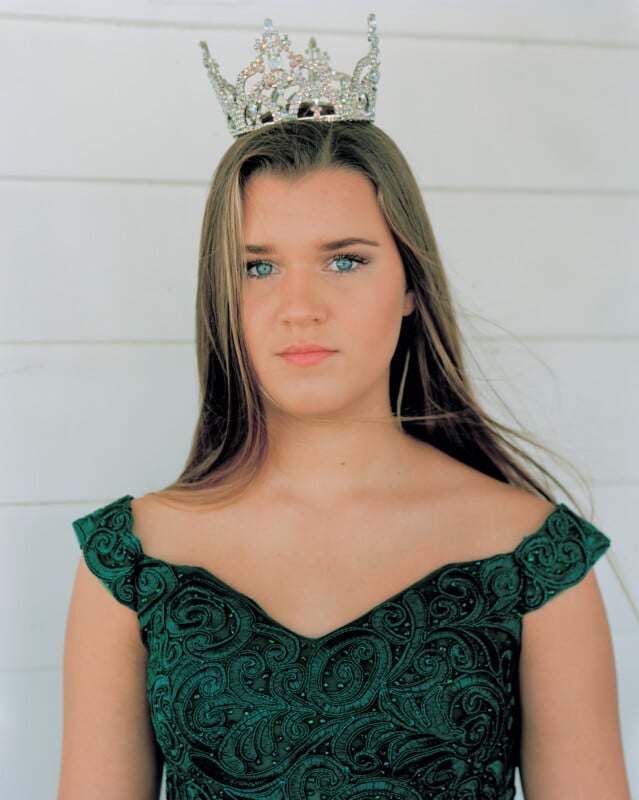 A young woman with long, straight brown hair wears a jeweled crown and a dark green, off-the-shoulder gown with intricate embroidery.