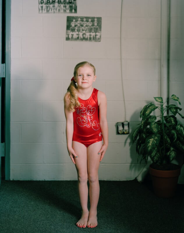 A young girl with blonde hair in a ponytail stands facing forward, wearing a red sequined leotard, in front of a white cinder block wall with two framed photos and a potted plant.