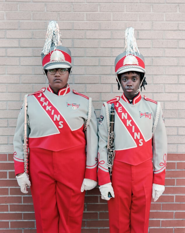 Two marching band members in grey and red uniforms stand side-by-side against a brick wall, holding a flute and a clarinet.
