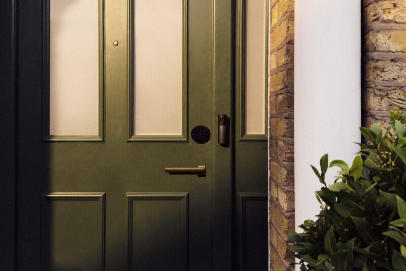 A dark green front door with frosted glass panels, a brass handle, a circular keypad lock, and a vertical smart doorbell, positioned next to a brick wall and a green plant.