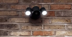 A black, dual-head outdoor security floodlight with a central camera lens mounted on a rustic brick wall.