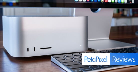 A silver Apple Mac Studio computer sits on a wooden desk next to a keyboard and a monitor stand, with a PetaPixel Reviews logo in the bottom right corner.