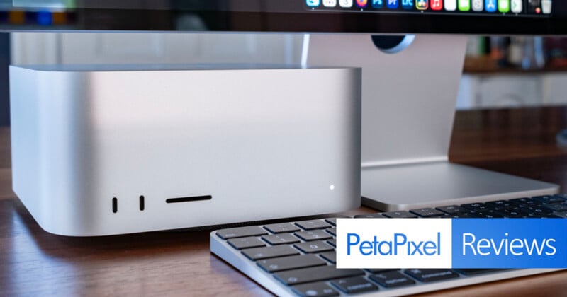 A silver Apple Mac Studio computer sits on a wooden desk next to a keyboard and a monitor stand, with a PetaPixel Reviews logo in the bottom right corner.