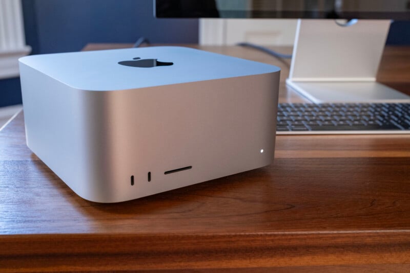 A silver Apple Mac Studio computer sits on a wooden desk next to a keyboard and a computer monitor.