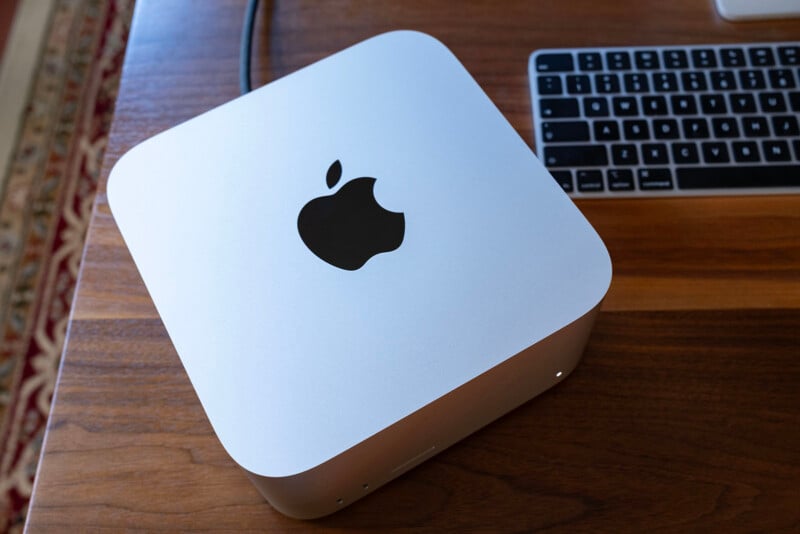 A silver Apple Mac mini sits on a wooden desk next to a wireless keyboard.