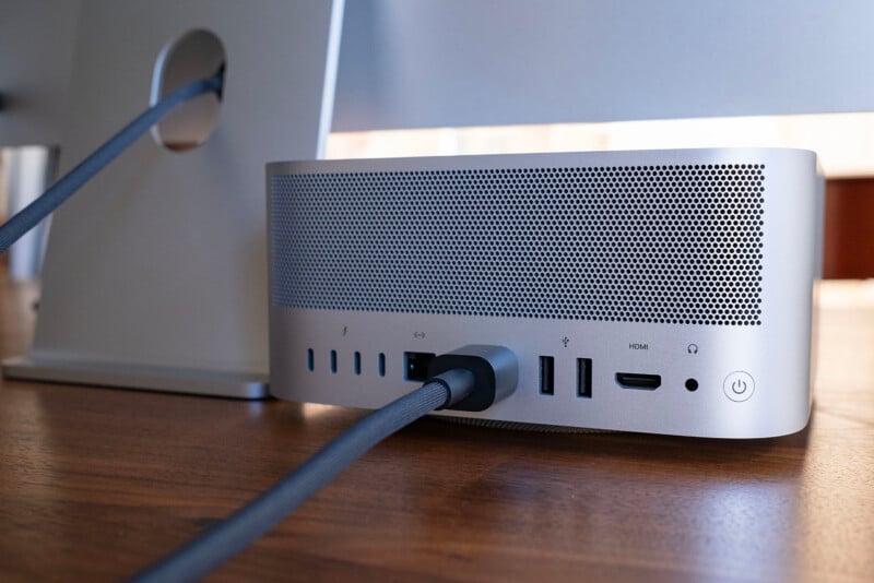 The back of a silver Mac Studio computer sits on a wooden desk, showing its various ports, a power cable, and a power button, with a monitor stand visible to the left.