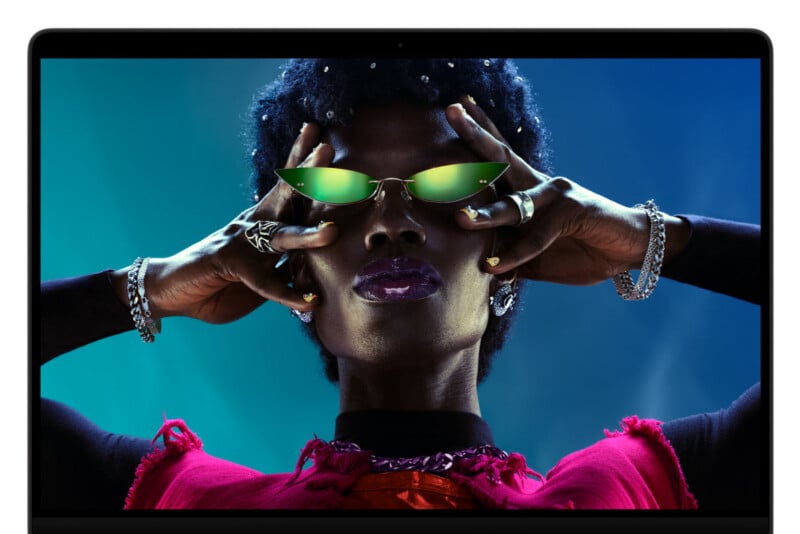 A person with dark skin and short, dark hair wears green-tinted cat-eye sunglasses, multiple rings, and bracelets while posing with their hands near their face against a blue background.