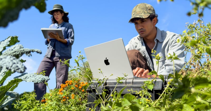 Two people in a field work on laptops, with a man in a camouflage cap in the foreground and a woman in a hat standing behind him.