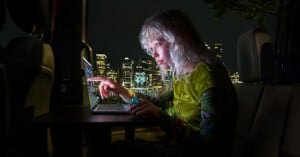 A person with blonde hair sits in the back of a vehicle at night, using a laptop while looking at a city skyline.