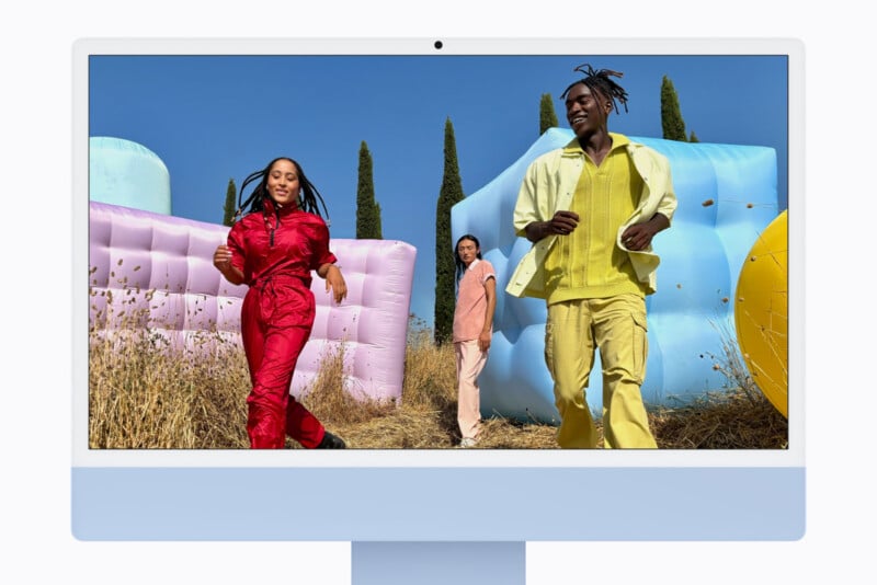 A light blue iMac displays a photo of three people walking through a field with large, colorful inflatable structures against a clear blue sky.
