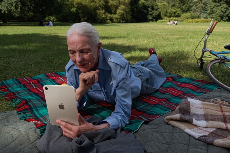 An older woman with white hair lies on a plaid blanket in a park, holding a tablet while looking at its screen.