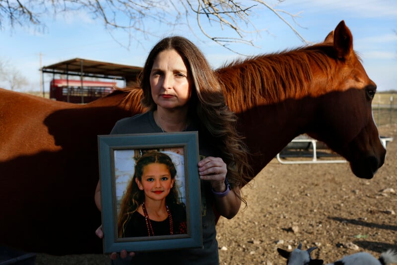 A woman with long brown hair stands outdoors holding a framed portrait of a young girl, with a brown horse standing behind her.