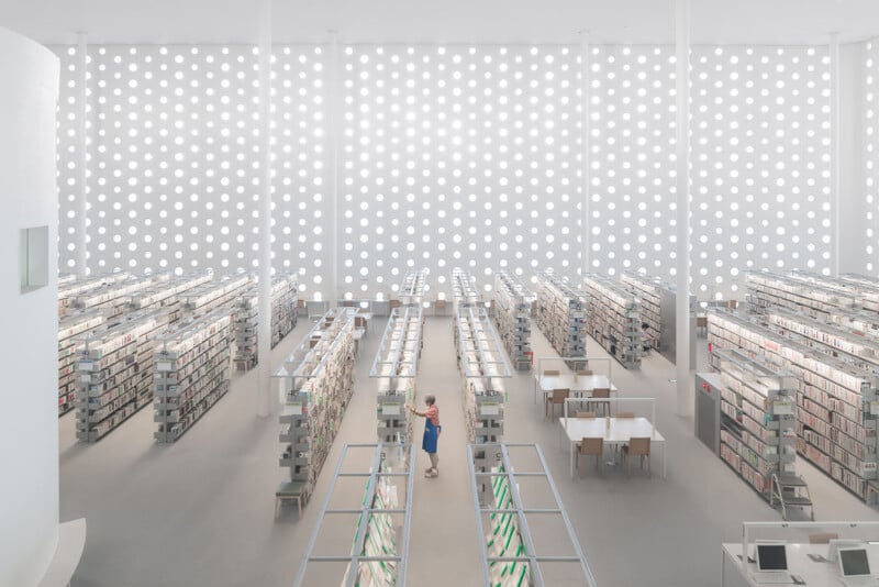 A high-angle view of a bright, minimalist library with rows of bookshelves, a person standing in an aisle, and a wall featuring a repeating pattern of circular cutouts.