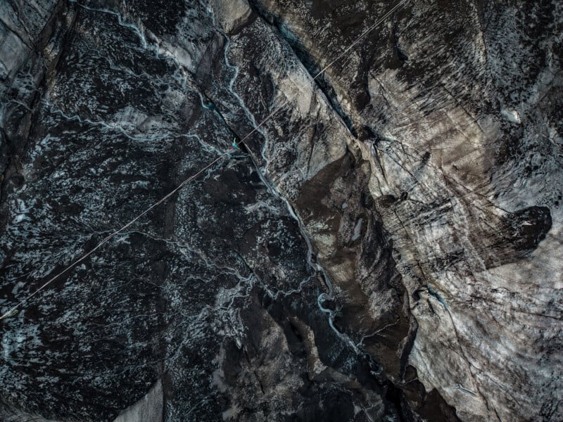 An aerial view shows a person walking on a highline stretched across a deep, dark, and rugged glacial crevasse.