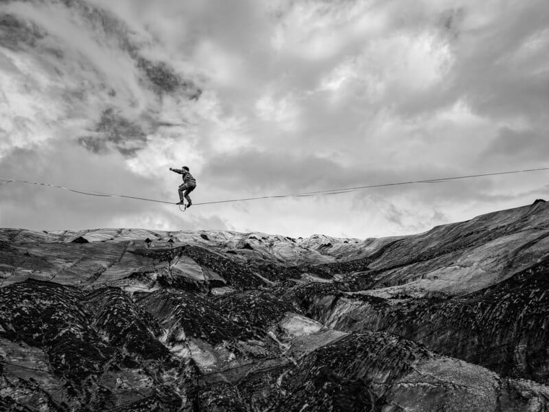 A person balances on a tightrope stretched across a vast, rugged, and rocky landscape under a cloudy sky.