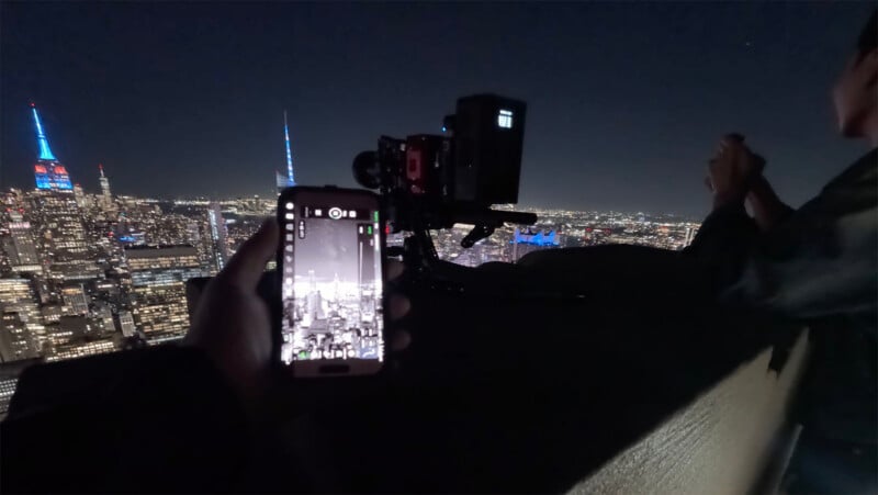 A person holds a smartphone displaying a camera interface while a professional cinema camera on a tripod captures a nighttime cityscape.