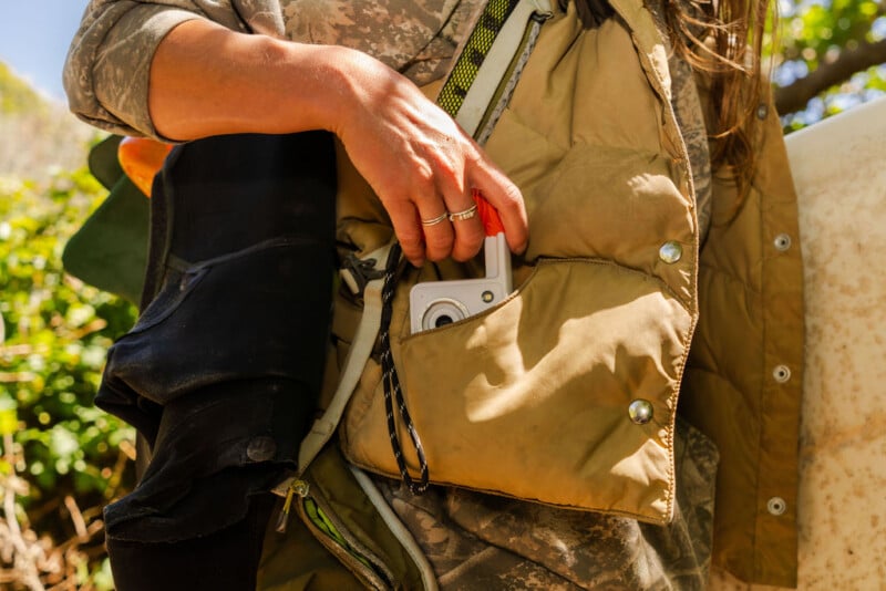 A person wearing a tan quilted vest and camouflage shirt places a small white camera into the vest's front pocket.