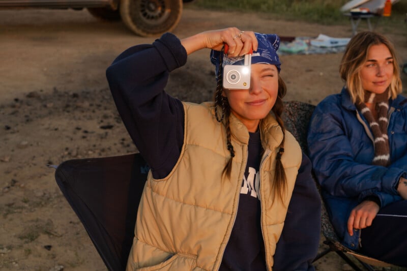 A woman wearing a blue bandana and tan vest holds a small white digital camera over one eye while sitting outdoors next to another woman.