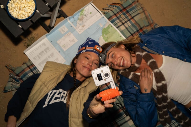 Two women lie on a plaid blanket outdoors, one holding a small camera toward the lens while a bowl of popcorn sits on a nearby table.