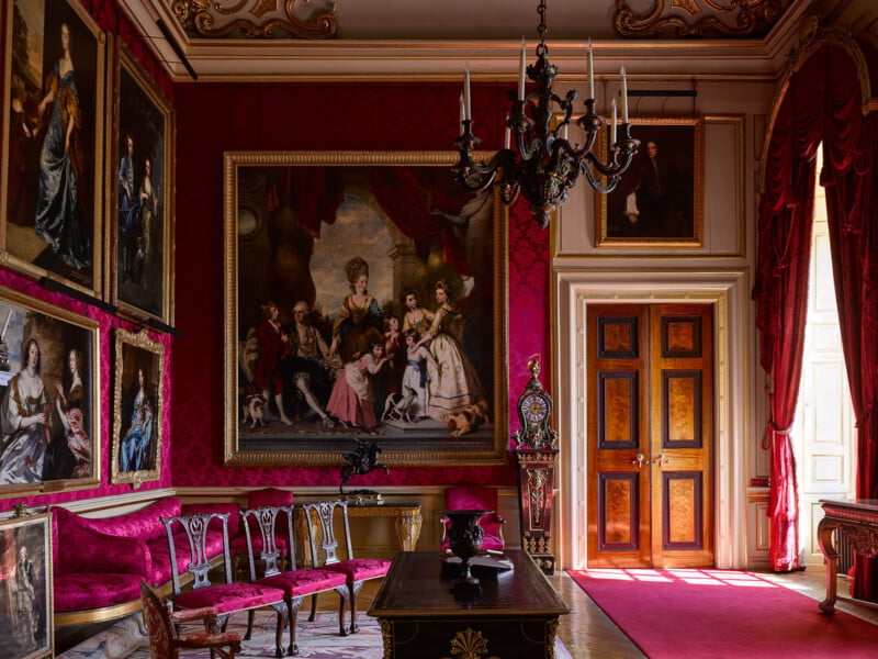 A grand room with red damask walls, a large central oil painting of a family, ornate furniture, a chandelier, and double wooden doors.