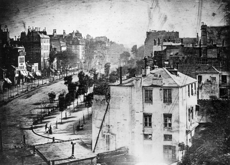 A black-and-white daguerreotype shows a quiet, empty Parisian street lined with trees and multi-story buildings, with a lone figure standing on the sidewalk.
