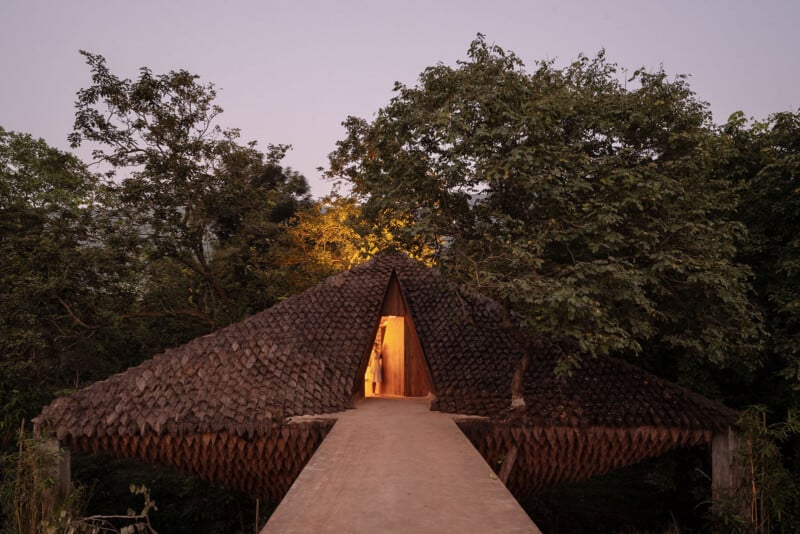 A person stands in the illuminated triangular doorway of a thatched-roof structure connected to a concrete walkway, surrounded by trees at dusk.