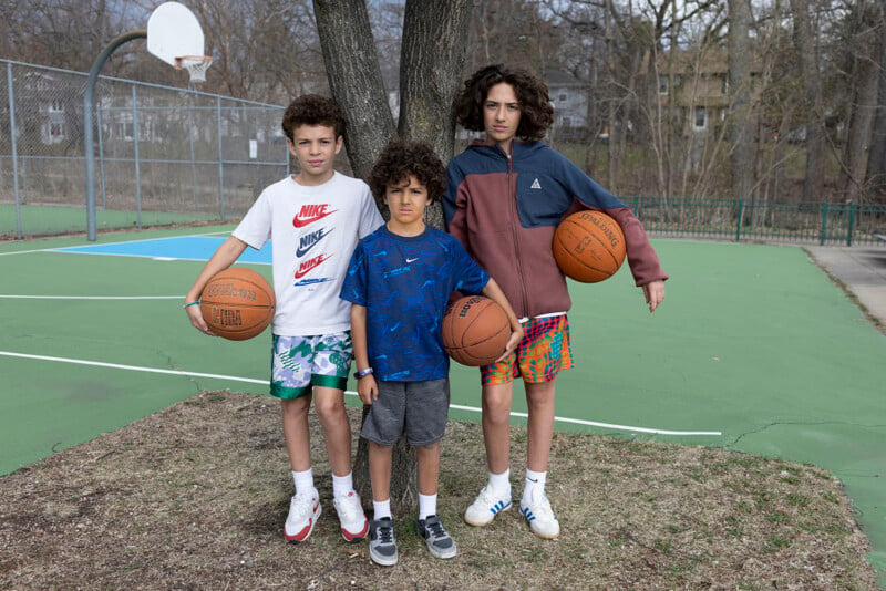 Three boys stand in a row in front of a tree on a basketball court, each holding a basketball.
