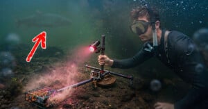 A diver underwater holds a camera rig with a light pointed at a submerged metal cage, while a large fish swims in the background.