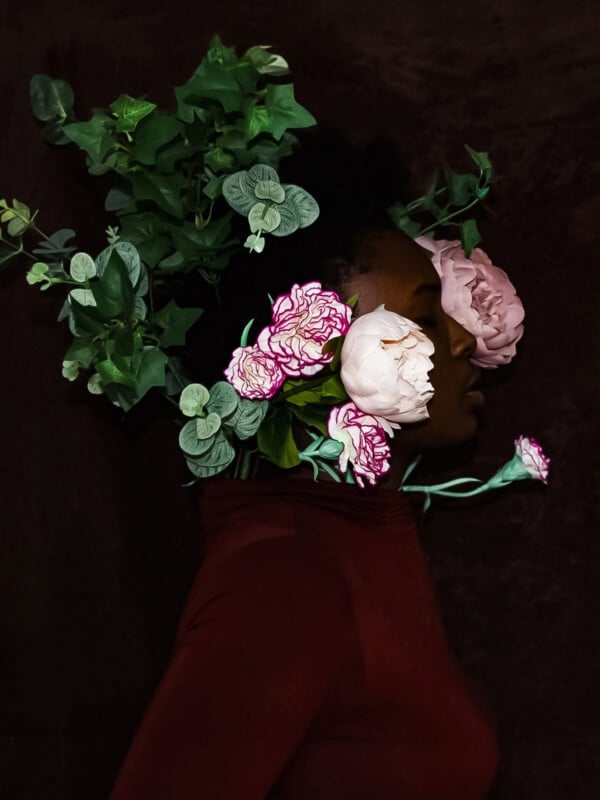 A person in profile wears a dark red top, their head and face partially obscured by an arrangement of green foliage and pink flowers.