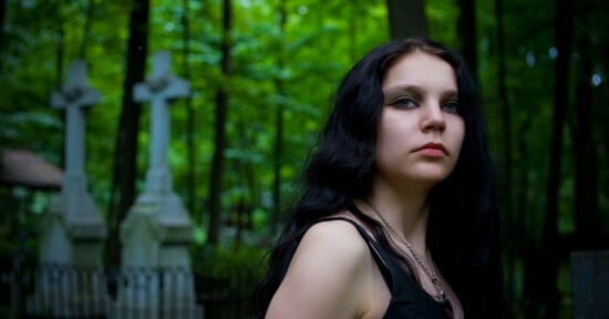 A young woman with long dark hair and dark eye makeup looks toward the camera, standing in a cemetery with stone crosses.
