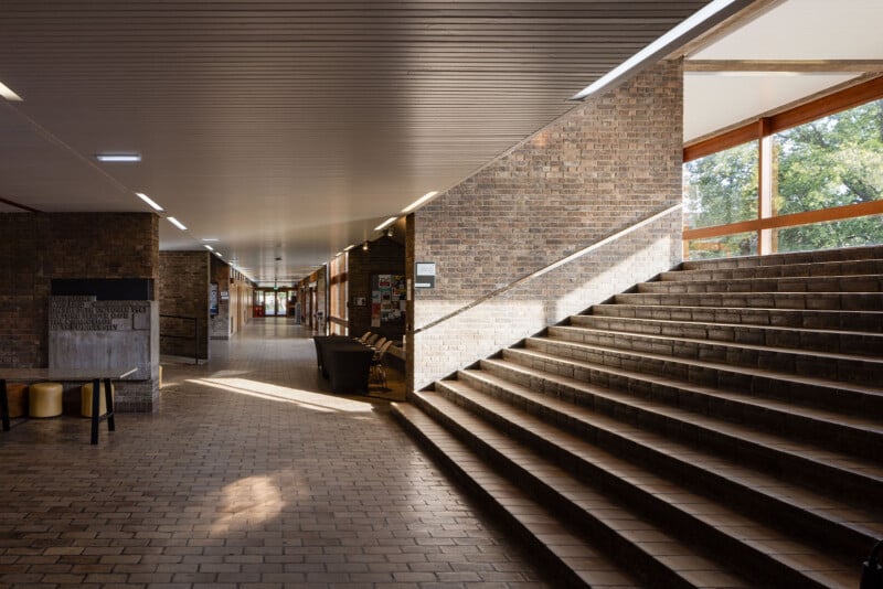 A wide, indoor corridor with brick walls and flooring features a large set of stairs on the right and a long, low ceiling with recessed lighting.