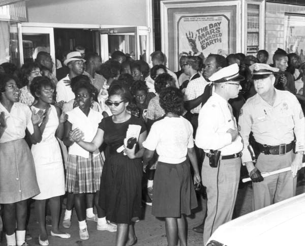 A group of young Black women stand in a line outside a theater, facing a police officer in uniform, with a movie poster for "The Day Mars Invaded Earth" visible in the background.