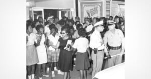A group of young Black people stand outside a theater entrance, facing two uniformed police officers who are holding a baton.