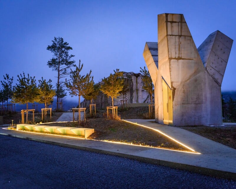 A modern concrete structure with a jagged, sculptural top stands next to a triangular paved area illuminated by embedded ground lights at dusk.