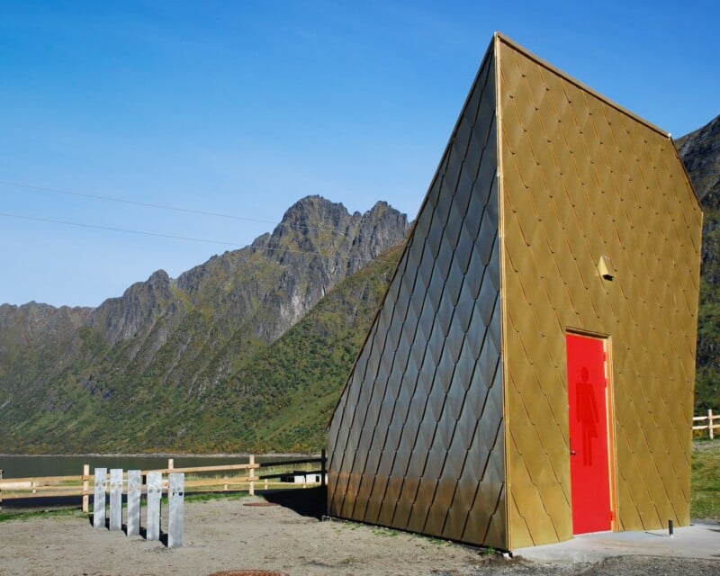 A modern, gold-tiled building with a bright red door featuring a female restroom symbol stands in front of a mountain range and a body of water.