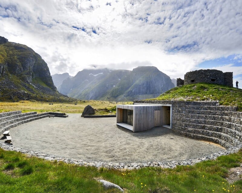 A modern wooden building with a flat roof sits in a gravel courtyard surrounded by tiered stone gabion walls, with a historic stone structure on a grassy hill and dramatic mountains in the background.