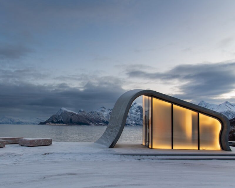 A modern, curved concrete structure with illuminated glass walls stands on a snowy shore overlooking a calm bay and snow-capped mountains.