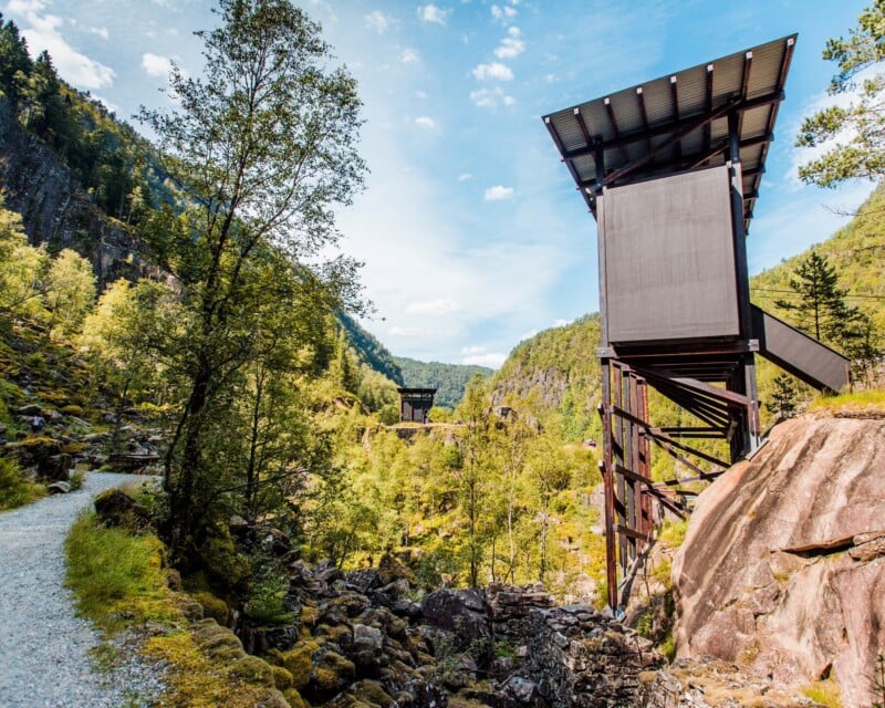 A gravel path leads through a rocky, forested valley toward a modern wooden observation tower perched on a hillside.
