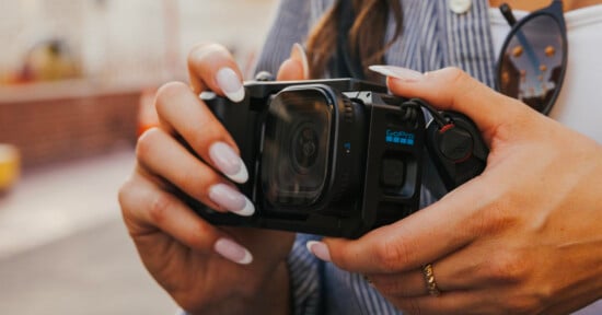 A person with long manicured nails holds a black GoPro camera in a protective housing, with a Peak Design anchor link attached to the side.