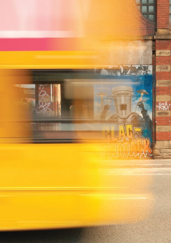 A motion-blurred yellow bus passes in front of a brick building featuring a mural of a bucket-headed figure with the text "CLAC ATTACK.