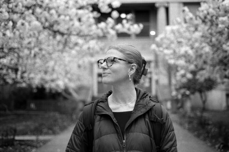 A person with glasses and hair pulled back looks to the side while standing outdoors between two flowering trees.