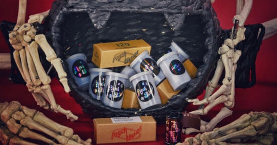 A black woven basket filled with Psych Blues film canisters and boxes is surrounded by skeletal hands on a red background.