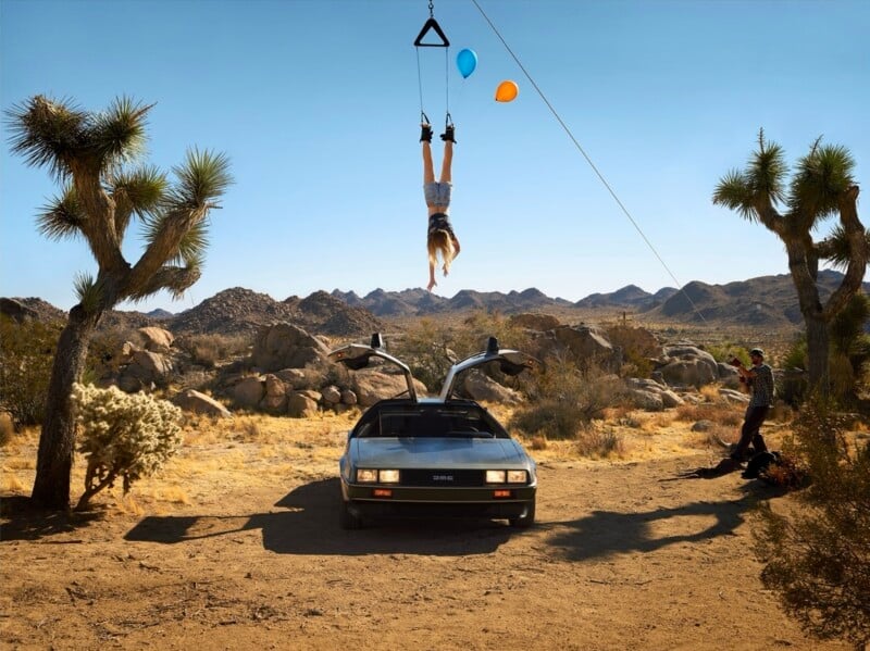 A person hangs upside down from a cable above a DeLorean car in a desert landscape, while a photographer stands to the side.