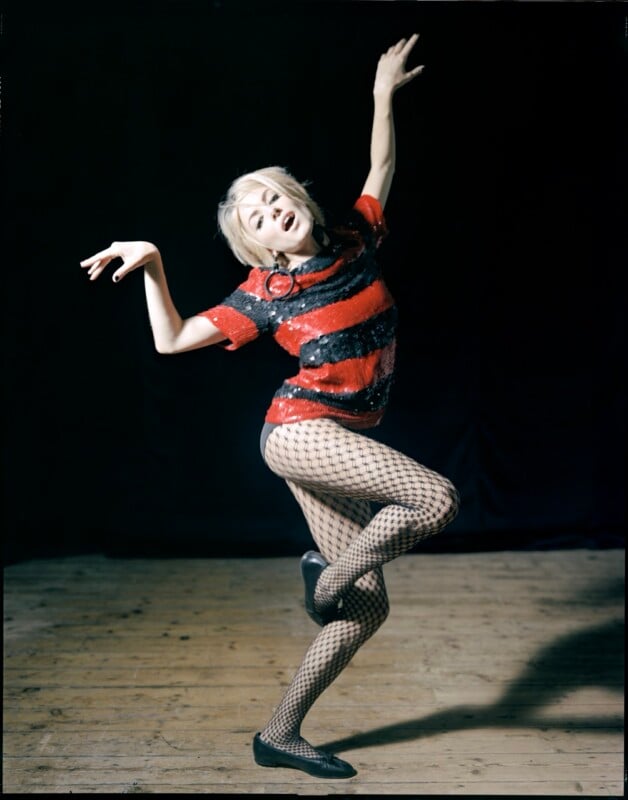 A blonde woman with a short haircut poses on a wooden stage, wearing a red and black striped sequined top, fishnet tights, and black ballet flats.