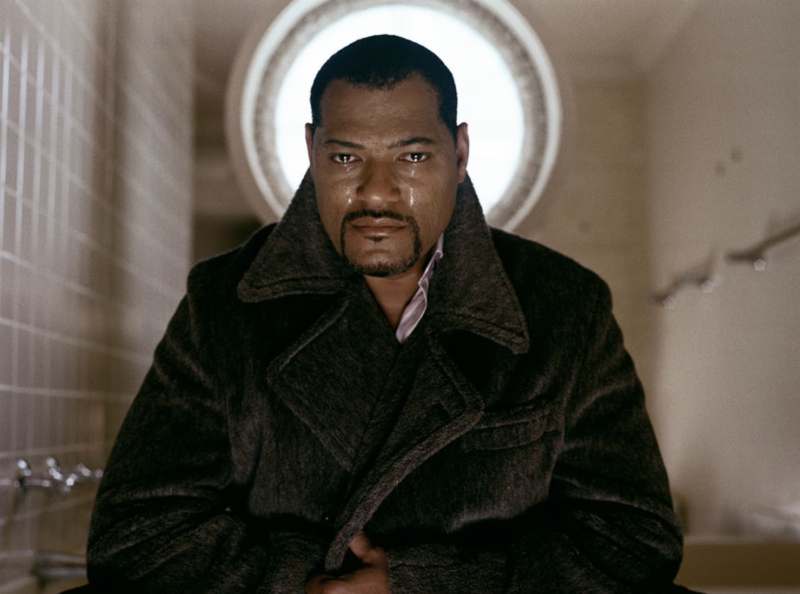 Laurence Fishburne with tears in his eyes, wearing a dark wool coat, stands centered in front of a circular window.