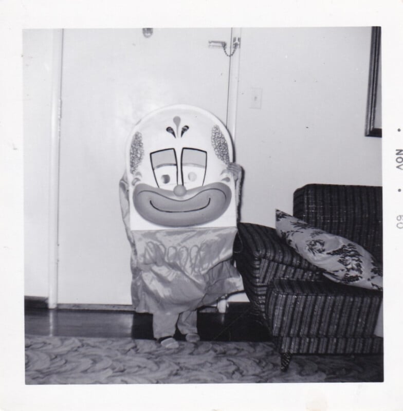 A child wearing a large, flat clown mask and costume stands in a living room next to a striped armchair.