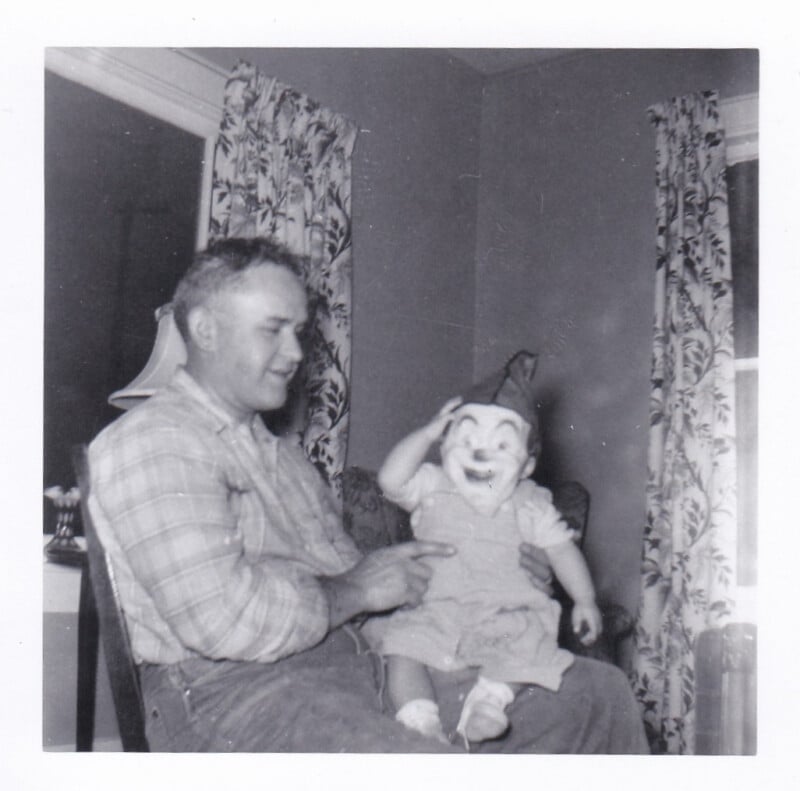 A man in a plaid shirt sits in a chair holding a baby wearing a clown mask and a pointed hat.